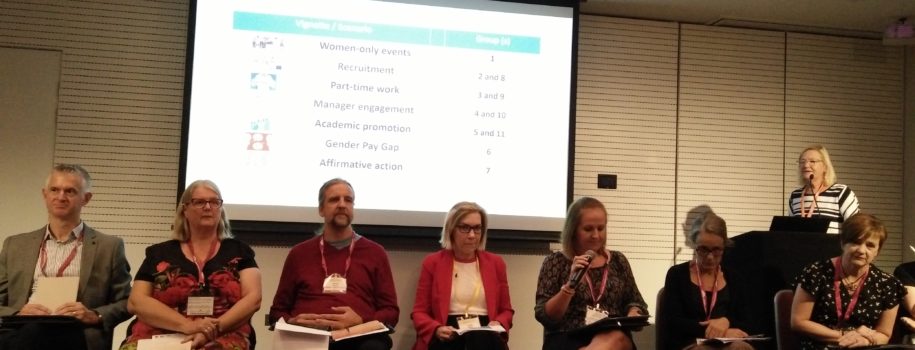 Near the front of a room, seven people (one speaking into a microphone) sit in a row of chairs. Behind them, a projector screen shows a list of vignettes/scenarios: women-only events, recruitment, part-time work, manager engagement, academic promotion, gender pay gap, affirmative action.