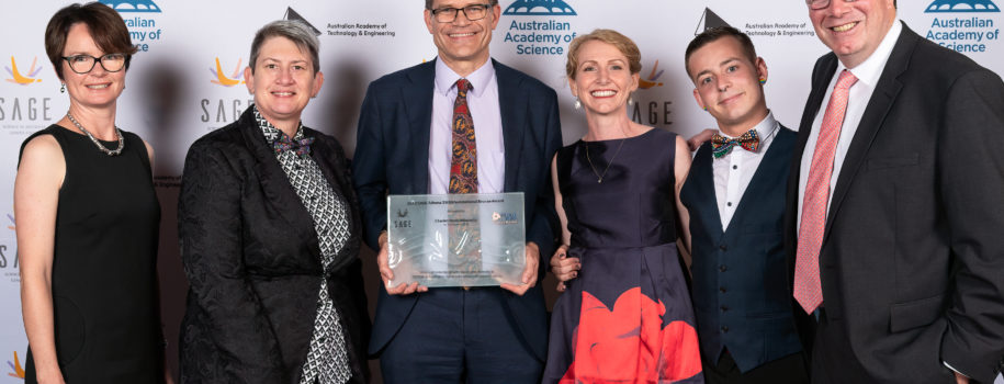 Six people in formal wear pose with a glass plaque in front of a large board adorned with SAGE, ATSE and Australian Academy of Science logos.