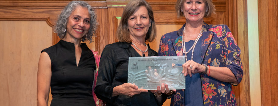 Three women pose for a picture on stage. One of them holds a glass plaque bearing the SAGE and Athena Swan Bronze Award logos.