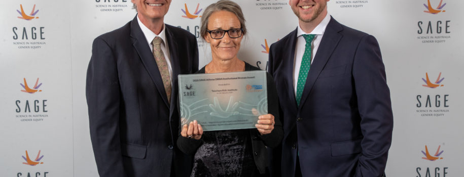 3 smiling people stand in front of a large board covered in SAGE logos. A woman in the middle is holding a glass plaque which reads “2020 SAGE Athena SWAN Institutional Bronze Award awarded to Telethon Kids Institute”.