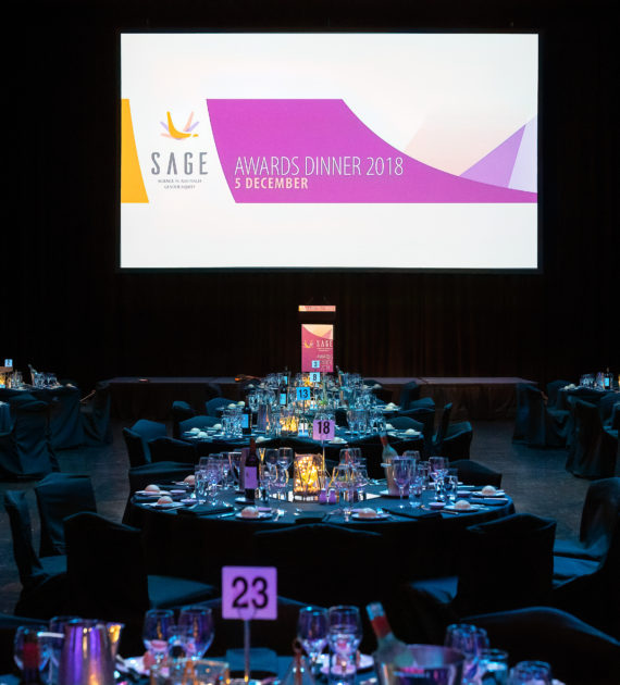A room with tables set for a formal dinner. A large screen at the front of the room says ‘SAGE Awards Dinner 2018, 5 December’.