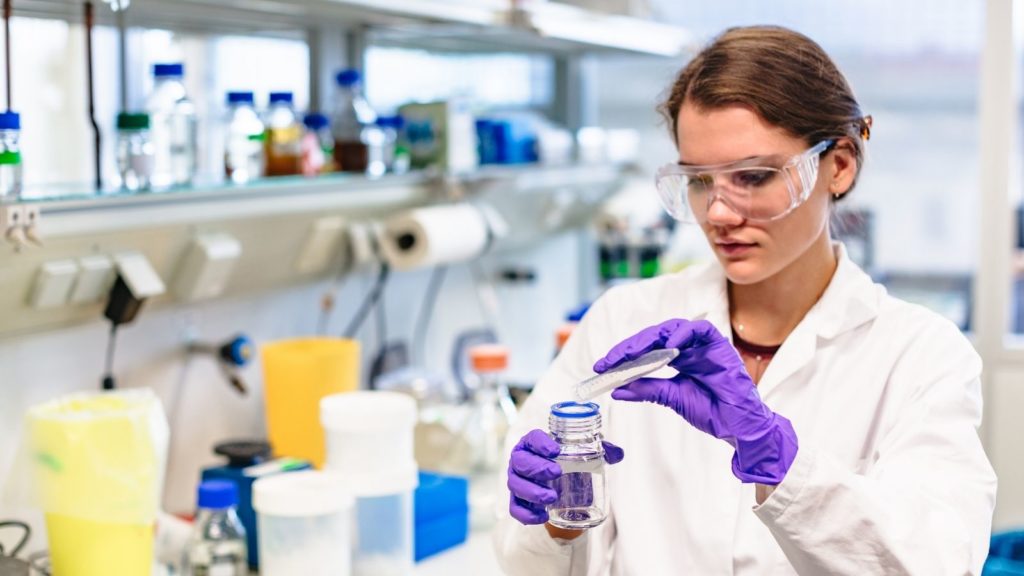 Woman works in a lab, wearing safety glasses, gloves and lab coat.