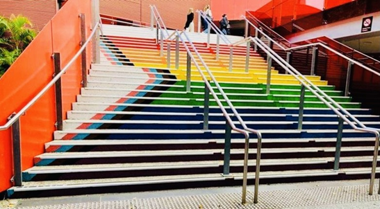 Outdoor staircase where the vertical face of each step has been painted to form a Progress Pride Flag when viewed from the bottom.