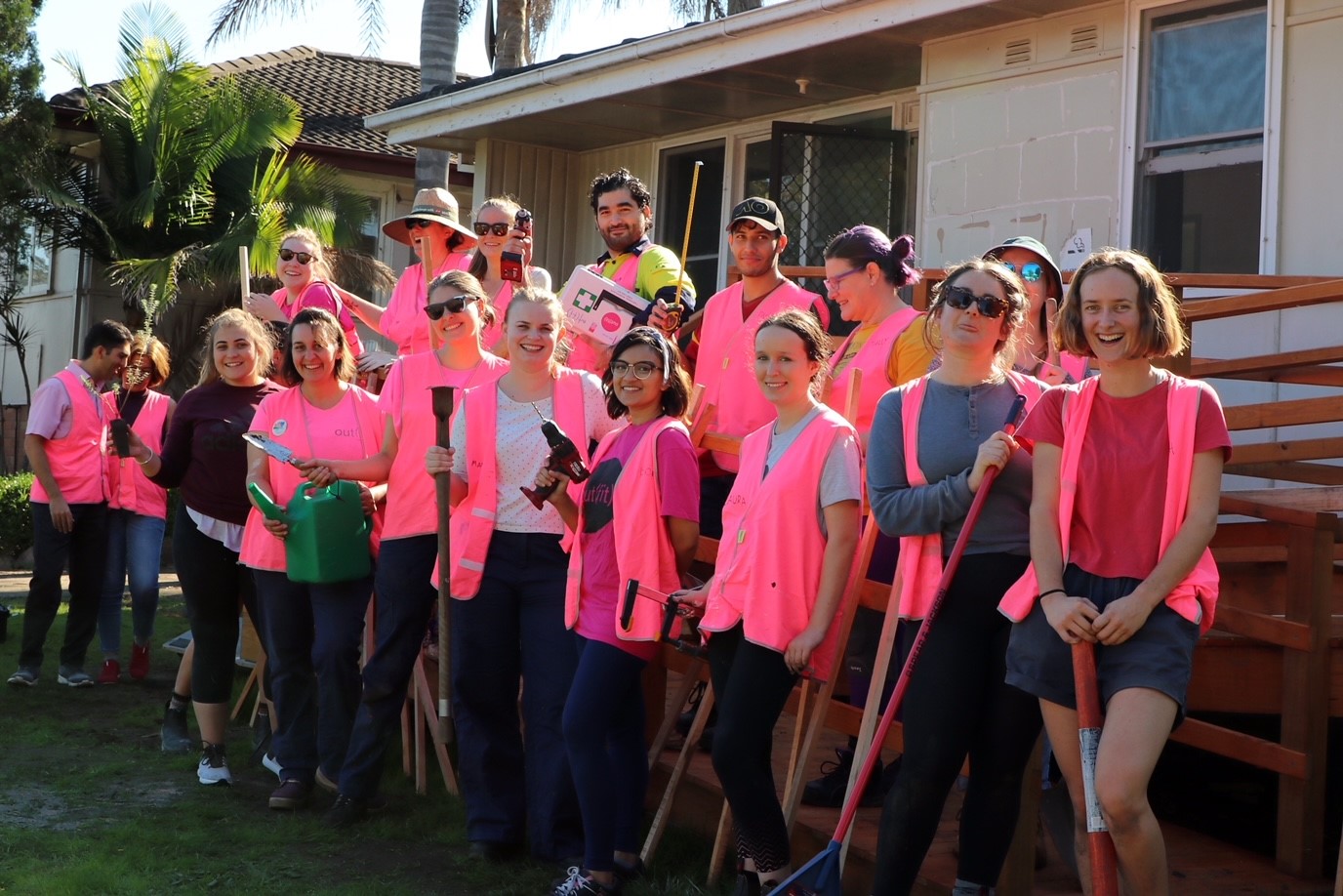 A group of people (mostly women) in a yard, wearing neon pink vests and holding power tools or gardening equipment.