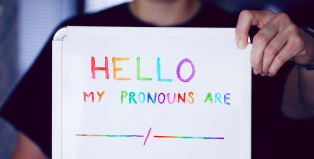 Person holding up a whiteboard that reads "Hello, my pronouns are" in rainbow-coloured ink.
