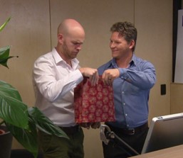 A smiling man hands a gift bag to another man, who is frowning uncertainly.